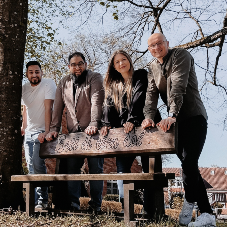 Pflegedienst Leer Vier Personen lehnen lässig an einer Bank unter einem Baum im Freien. Plattdeutsch